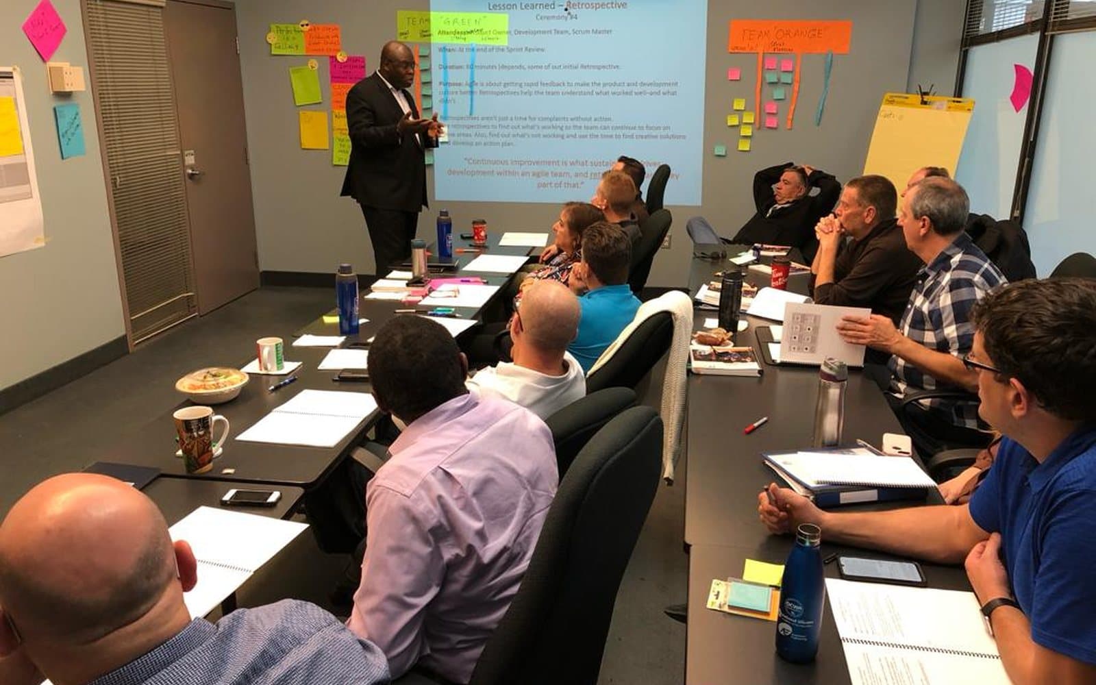 One of our trainers presenting a sprint retrospective to a seated group, with two teams' sticky-note walls behind him.