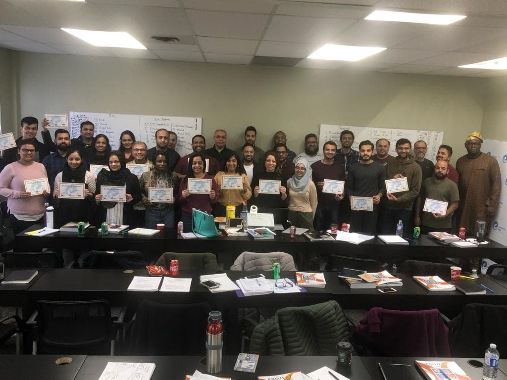 A large cohort standing at the front of a training room, each person holding a printed course certificate, with project management workbooks on the desks.