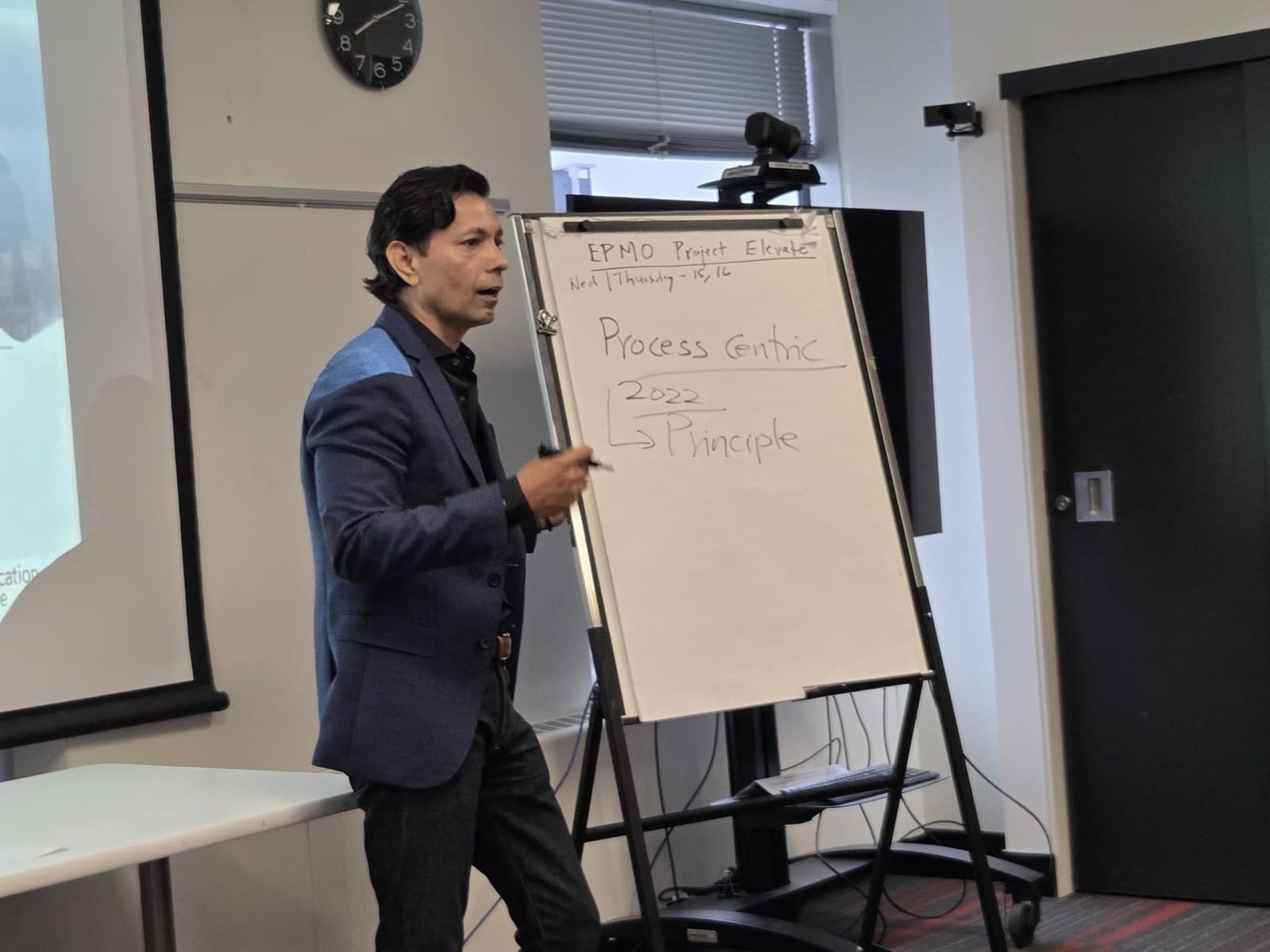 Hemant Dhariyal in a blue blazer, marker in hand, mid-sentence beside a flipchart in a meeting room.