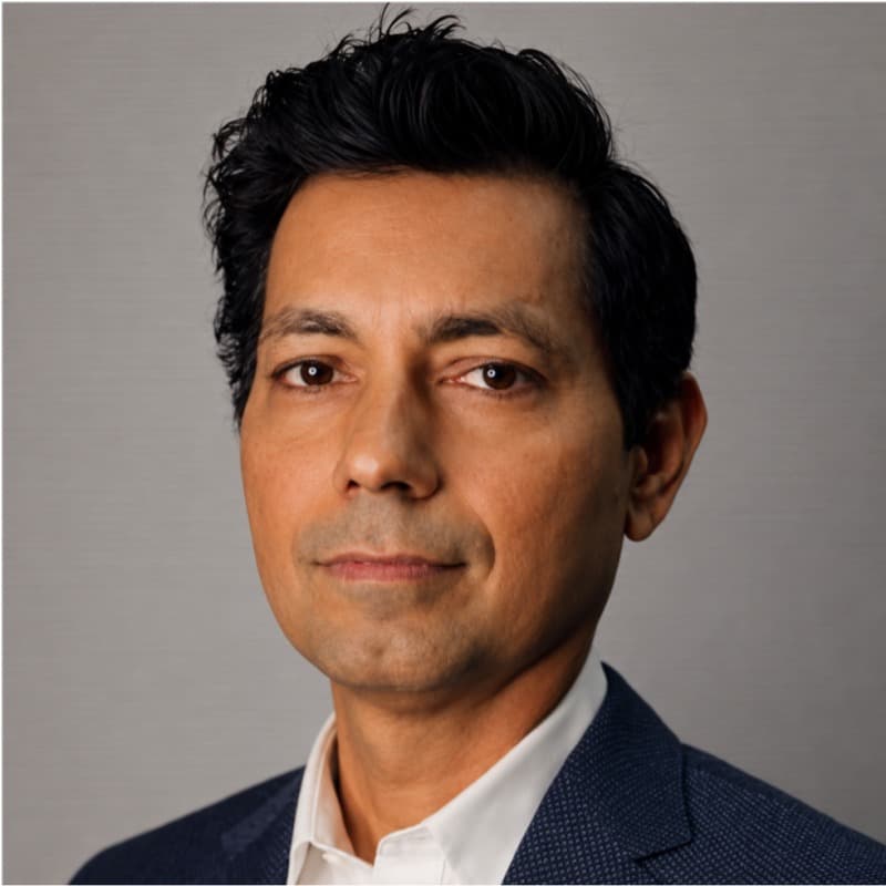 Hemant Dhariyal, in a navy blazer and open-collar white shirt, photographed against a plain grey studio background.