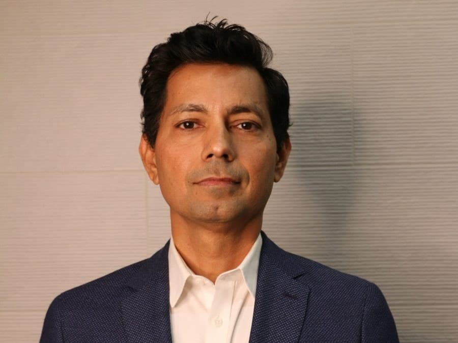 Hemant Dhariyal in a navy blazer and white open-collar shirt, standing against a pale wall.