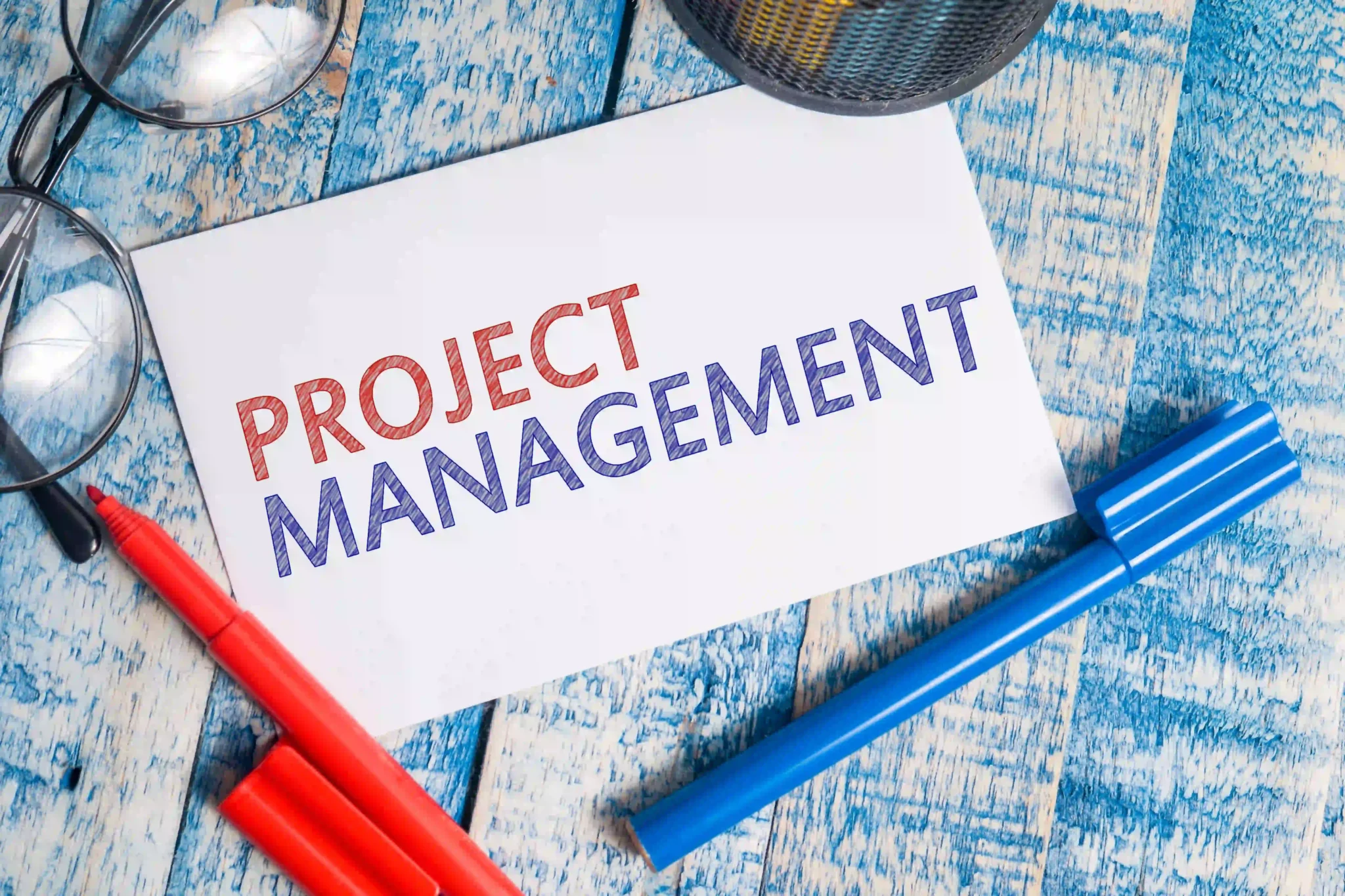 Project management written on a white note card surrounded by markers and eyeglasses on a wooden table.