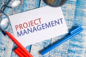 Project management written on a white note card surrounded by markers and eyeglasses on a wooden table.
